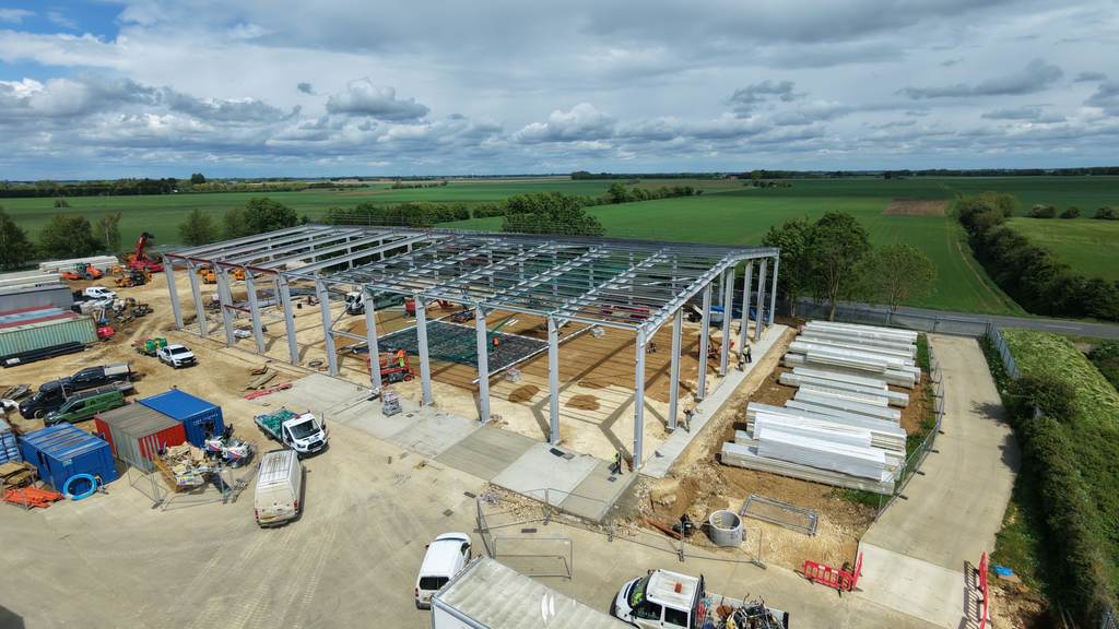 Aerial drone view of a steel portal-frame building under construction in open farmland near Peterborough, with the site compound, cabins and stacked materials alongside