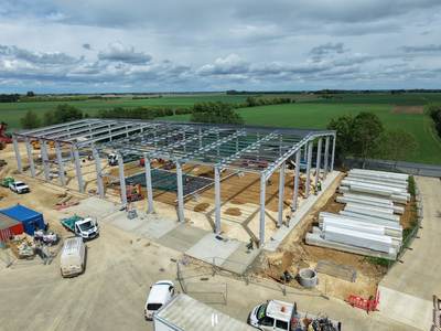 Aerial view of the steel portal frame under construction in open farmland near Peterborough, with the site compound and stacked materials alongside