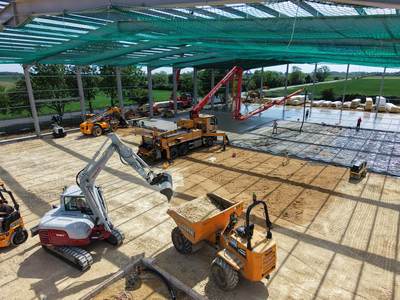 Drone view inside the frame: a concrete pump boom pours the slab over steel mesh while an excavator, roller and dumper prepare the sub-base