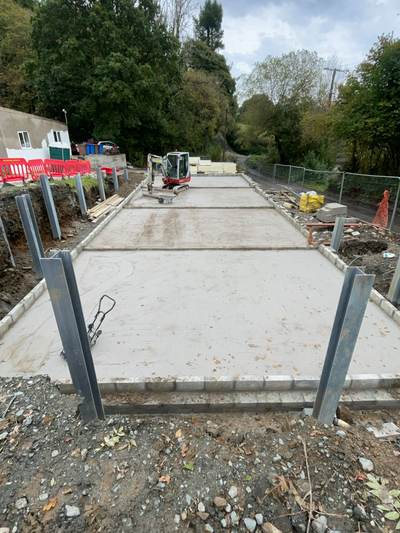 Completed concrete slab bays with kerb edging and the steel posts of a new retaining wall set in place, a mini digger working behind